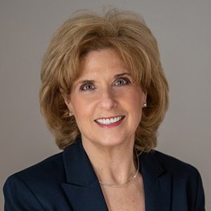 A woman with short, light brown hair smiles at the camera. She is wearing a dark blazer, a light top, and a thin necklace, set against a plain, neutral background.