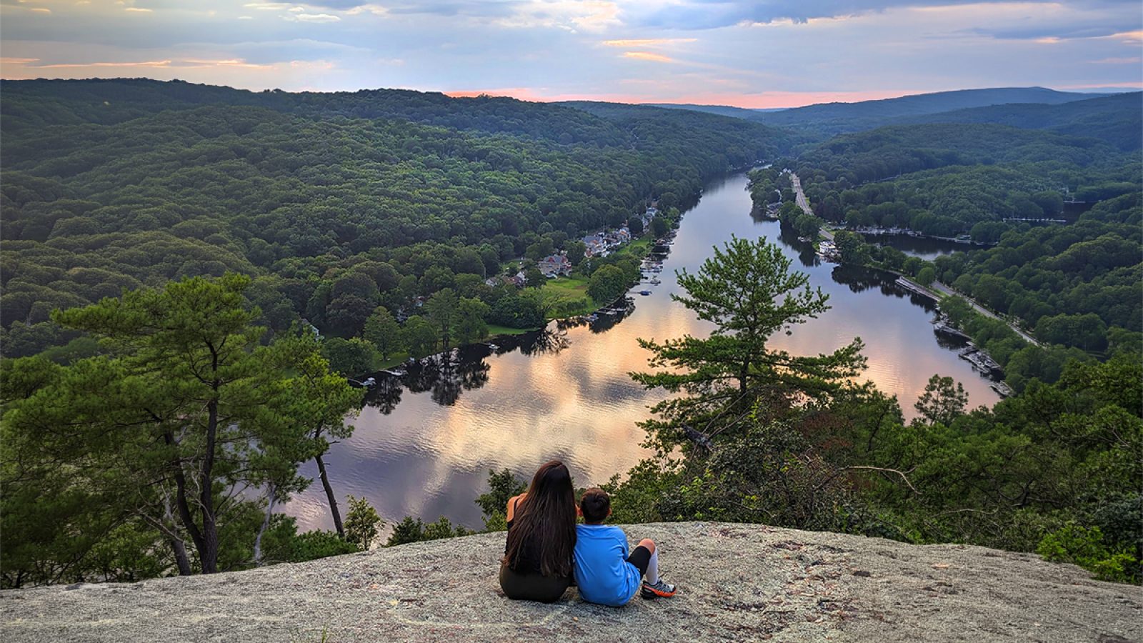 A woman and a child sit on a rocky ledge overlooking a river winding through a lush, green forest at sunset, with the sky reflecting on the water.